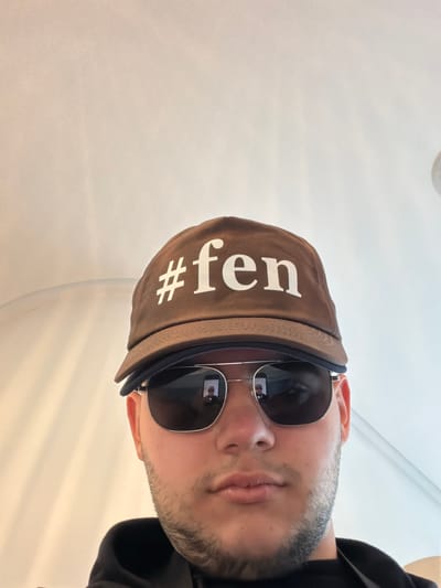 The image presents a close-up, chest-up view of an adult male. The individual wears a brown baseball cap with the white text "#fen" across the front, and dark-lensed aviator-style sunglasses with silver frames. The sunglasses reflect a smartphone held by the subject, suggesting a self-portrait. A light beard or stubble covers the chin and cheeks. The subject is wearing a dark-colored garment. The background is an illuminated, light-colored indoor setting, featuring a subtle curved wall or structure on the left and a faint circular fixture in the upper right. The specific location in Grau i Platja, Spain, cannot be determined from the visual information provided.
