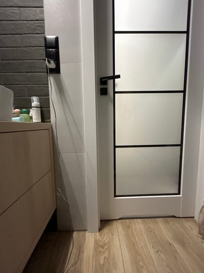 The image depicts an interior scene, likely a bathroom or hallway, with a wooden floor and a white door with frosted glass panels. To the left, a portion of a light-colored cabinet with drawers is visible, topped with a few toiletries, including a white dispenser bottle and a few small round objects. Adjacent to the cabinet, a white tiled wall features a black electrical outlet with a black and white checkered cord plugged in. The overall impression is one of a modern, domestic setting. The location in Siedlce, Poland, is not discernible from the image itself.