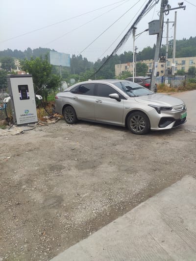 In Jianyang City, China, a silver sedan is parked at a public electric vehicle charging station. The charging station, labeled with Chinese text and a 160kW power rating, is connected to the car's charging port. The surrounding area is a dirt and gravel lot, indicative of an outdoor public space. In the background, a street, utility poles with numerous wires, and buildings are visible. The sky is overcast, suggesting a cloudy day.