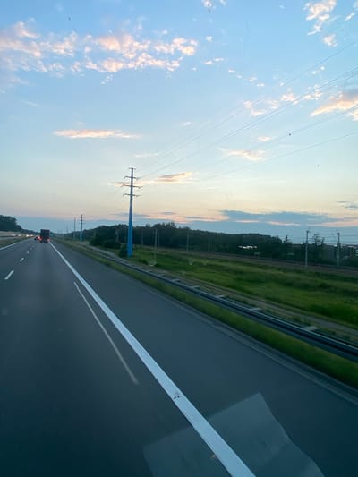 The media file depicts a road trip perspective captured from a vehicle, likely a car, traveling along a highway in Kokotów, Poland. The scene is oriented vertically, suggesting the image was taken while the vehicle was in motion. The main subjects are the highway itself, flanked by green grassy areas and a line of trees on the left, and electrical power lines stretching across the sky on the right.
The highway is asphalt, marked with white lines, and appears to be multi-lane. In the distance, a dark-colored truck is visible, with its brake lights illuminated, suggesting it is moving ahead. Other vehicles are faintly visible further down the road. The sky is a pale blue with scattered clouds, tinged with orange and pink hues, indicating it is either sunrise or sunset. The presence of electrical pylons, characteristic of power transmission infrastructure, reinforces the setting of a highway in a developed area.
The weather appears clear and calm. The overall mood is one of transit and perhaps contemplation, typical of a journey on a highway. There is no visible text in the image. The time of day is likely either early morning or late evening due to the sky's coloration. The location is specified as Kokotów, Poland.