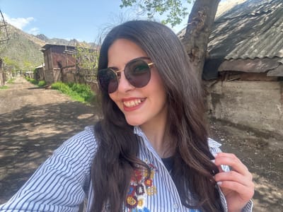 A woman wearing sunglasses and a striped shirt with floral embroidery poses in Agarak, Armenia. The background features a rustic, weathered building, possibly a shed or outbuilding, with a corrugated roof. In the distance, there are hills or mountains and a clear blue sky. The immediate surroundings seem to be a rural or semi-rural setting with foliage and ground textures visible.