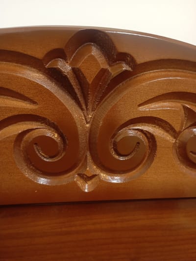 The image is a close-up shot of intricately carved dark brown wood. The carving features repeating scrollwork and floral motifs, with a polished finish that reflects light. The wood appears to be part of a larger decorative element, possibly furniture or an architectural feature. The background is a plain, light-colored surface, suggesting an indoor setting. The image does not contain any information about its location in the city of Ararat, Armenia.