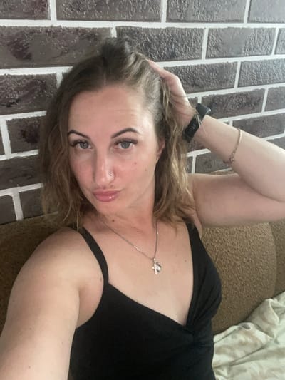 A woman is lying on her side, facing the camera. She has light brown hair that falls around her shoulders. She is wearing a black top with a thin strap over her shoulder and a necklace with a small charm. Her right arm is bent at the elbow, with her hand resting on the back of her head. She has a bracelet on her left wrist and a black watch. Her eyes are open and looking at the camera. She has a nose piercing.
The background appears to be a tiled floor with a brown, textured surface to the right. The tiling looks like dark brown bricks with white grout. The overall lighting suggests it is daytime, though the indoor setting makes it difficult to determine natural light conditions. There are no visible cues for weather. The image is likely taken in a residential setting. The woman's expression appears neutral or relaxed. No text is visible in the image.