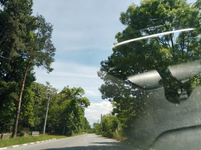 The image captures a view from a vehicle traveling on a paved road lined with lush green trees. The road has a white dashed line on the left edge. On the left side of the road, there is a brown wooden pole with wires running overhead, and beyond that, a stone wall with some vegetation. On the right side, there are more dense trees, and a faint reflection of the interior of the vehicle is visible on the windshield. The sky is a clear blue with a few wispy clouds. The scene appears to be in a rural or suburban area. The specific location in Mirovo, Bulgaria, is indicated by the user.