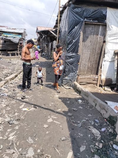 The image depicts a street scene in Oworonshoki, Nigeria, under bright daylight with a partly cloudy sky. The ground consists of an unpaved, uneven dirt path heavily littered with refuse, including plastic fragments, paper, and debris. A low concrete curb runs along the right side of the path, bordering a drainage channel also filled with waste.
In the foreground, three individuals are present. On the left, a shirtless male with dark, curly hair wears dark trousers and light-colored sandals; his light-colored underpants are visible above his waistband. He stands, looking down at an object held in his hands. To his right and slightly forward, a young child, possibly a toddler, stands facing left, wearing a light-colored shirt, patterned shorts, and small shoes. Further to the right and slightly behind the child, a female walks forward (towards the right), wearing a dark sleeveless top, a patterned wrap skirt over dark leggings or trousers, and sandals. She holds a light-colored shoulder bag and has an object held to her mouth.
The environment is characterized by densely packed, rudimentary structures. On the left side of the path, multiple wooden shacks with corrugated metal roofs are visible, some appearing to be constructed from scrap materials, one with a faded light blue section. On the right side, a structure is predominantly constructed from wooden planks and extensively covered with black plastic sheeting (tarpaulin), featuring a rough wooden door. Overhead wires span across the scene. Further down the path, additional indistinct figures and structures are visible, including one person in a pink or purple garment. Shadows indicate the sun is high in the sky, casting shadows towards the right.