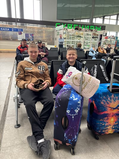 Two boys are sitting in an airport waiting area. They are seated on chairs with metal frames and gray seats. Behind them are several pieces of luggage, including two brightly colored suitcases and a white shopping bag with Arabic script. In the background, there are several other people waiting, along with retail shops selling jewelry and souvenirs. The signage is in Russian, with "KACCA" visible. It is unclear if they are in Boldino, Russia, as there are no identifying features.