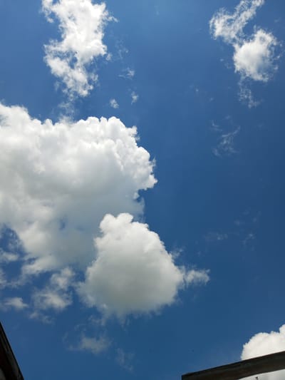 This is a photograph of a bright blue sky with scattered white clouds. The clouds are fluffy and appear to be cumulus clouds. In the lower left and lower right corners of the image, there are parts of man-made structures, likely wooden beams or eaves of a building, indicating an outdoor scene. There are also a few small dark specks in the sky that could be birds. The image appears to have been taken during the daytime, with the sun likely high in the sky given the brightness and the contrast of the clouds. The location is identified as Zavyalovo, Russia.