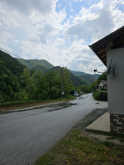 The image presents an uphill road in the village of Erma Reka, Bulgaria, on a wet day. The asphalt road is slick with water. Verdant mountains covered with dense forest form the backdrop. Overcast sky. The image is taken from under the roof of a building that is on the right side of the road. The building has a satellite dish on its side.