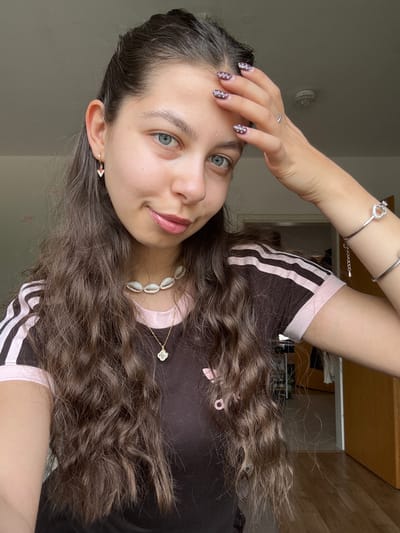A young woman with long, wavy brown hair is posing for a selfie indoors. She is wearing a dark brown Adidas-style shirt with light pink stripes and trim. She has on jewelry, including a shell necklace, a pendant necklace, a silver bracelet with a heart charm, and earrings. Her nails are painted dark with white dots. Her right hand is raised, with fingers touching her forehead. Her expression is serene.
The background is partially obscured, showing a doorway leading to another room with light-colored walls and flooring. An overhead light fixture is visible on the ceiling behind her. The location is likely an interior space in Bruchköbel, Germany.