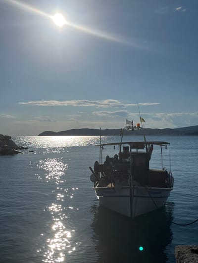The image depicts a serene harbor scene in Potos, Greece, on a clear, sunny day. The sun, positioned high and slightly to the left, casts brilliant reflections across the calm, blue water. A fishing boat is anchored in the foreground, its silhouette dark against the bright water. The boat has a cabin structure and appears equipped with fishing gear. Two flags, one of which is the Greek flag, are visible on the boat's mast. In the distance, rolling hills form the coastline, with a town visible along the shore. On the left side of the frame, a rocky breakwater extends into the water. The overall atmosphere is one of tranquility and coastal life.