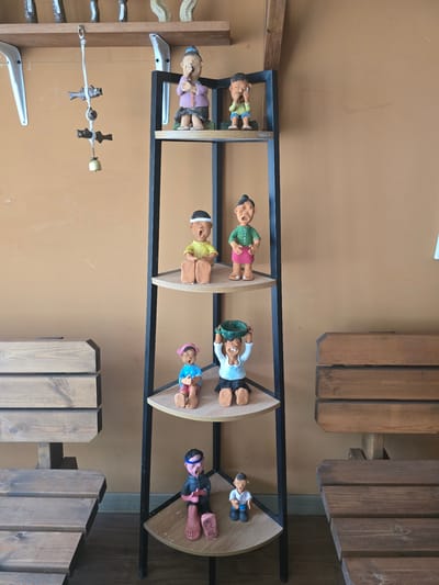 This media file captures a display of handcrafted figurines arranged on a multi-tiered shelf unit. The figurines appear to be made of clay and depict stylized human figures, some with exaggerated features and cartoonish expressions. Each figurine is dressed in colorful clothing, including green shirts, pink skirts, blue shorts, and light blue shirts. Some figures are holding objects, such as a green basket or what appears to be a toy.

The shelves are circular and made of wood, supported by a black metal frame. The backdrop is a plain, light brown wall. In the background, wooden table and chair elements are visible, suggesting the display might be in an outdoor seating area or a cafe. The overall presentation evokes a whimsical and playful atmosphere.

There are no discernible signs of time of day or weather conditions. The figurines themselves do not convey specific emotions beyond a general sense of liveliness. No text is visible on the figurines or within the immediate display area. The context provided places this scene in Gumi-si, South Korea, hinting at a local craft or artistic display.