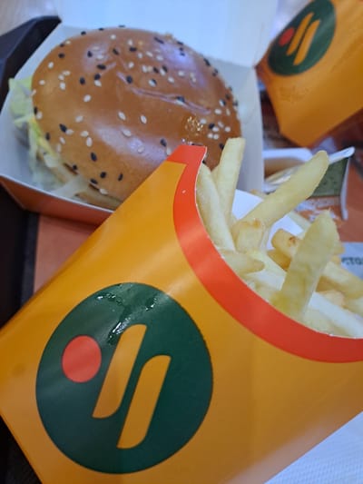 A close-up shot presents a burger and a container of french fries on a dark surface. The burger is in a white cardboard box, revealing a sesame seed bun. The french fries are in an orange cardboard container with a green and orange logo. The photograph appears to have been taken in a location of City of Yegoryevsk, Russia.