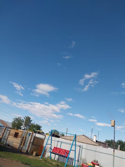This is a photograph taken outdoors in Трудовое, Kazakhstan. The image is oriented vertically. A bright blue sky with scattered white clouds dominates the left side of the frame. The right side of the image shows a residential area with several structures. In the foreground, a blue metal swing set with a red seat is visible. Behind the swing set, there are buildings constructed with corrugated metal and wood. One building appears to be a shed or small workshop with a wooden facade and a small window. Further back, a larger building with a corrugated metal roof and siding is present. Several trees with green foliage are scattered throughout the scene, suggesting it is daytime and likely during a warmer season. There are some colorful tires stacked near the fence of one of the buildings. A bird feeder or nesting box is mounted on a metal pole in the upper right portion of the image. The scene appears to be a residential yard or backyard. No people are visible in the image, and there is no discernible text. The lighting suggests it is daytime, likely morning or afternoon, with clear and sunny weather.