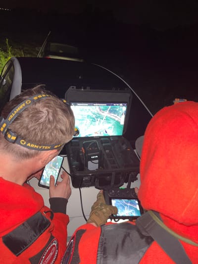 Two individuals, wearing matching red hooded jackets and headlamps, are engaged in operating a drone at night. One individual on the left is holding a smartphone displaying a map interface, while the other individual on the right is operating a drone controller with a screen. They are positioned next to a black vehicle, and a case containing drone equipment is open between them. The scene appears to be outdoors, with vegetation visible in the background, and is consistent with a location in or around the village of Badashka, Russia. The lighting suggests it is dark, with the headlamps and screens being primary light sources.