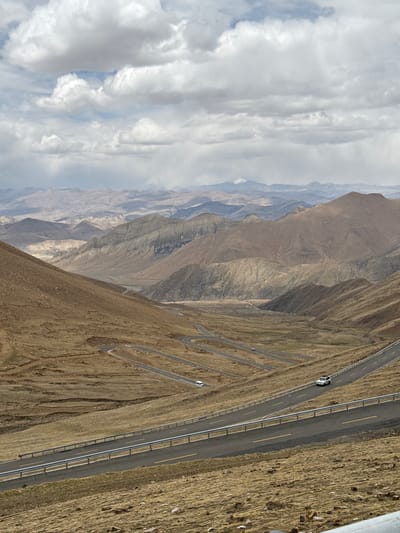The image depicts a winding mountain road in the vicinity of Zagor town, China. The road, with guardrails, traverses a steep, arid mountain landscape. There are cars on the road. The sky is overcast with a mix of clouds. The mountainous terrain consists of layered, rocky slopes with sparse vegetation.
