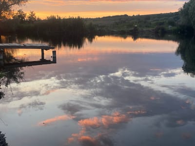 The image depicts a serene lake scene at sunset in Cotihana, Moldova. The sky is ablaze with hues of orange and pink, reflecting beautifully on the calm water. A wooden jetty juts out into the lake from the left, its weathered planks creating geometric shapes. Tall reeds line the far bank, interspersed with lush green trees and rolling hills in the background. The water's surface mirrors the sky and the surrounding vegetation, creating a dreamlike, symmetrical composition. There are no people or animals visible in the frame. The overall atmosphere is one of tranquility and natural beauty.