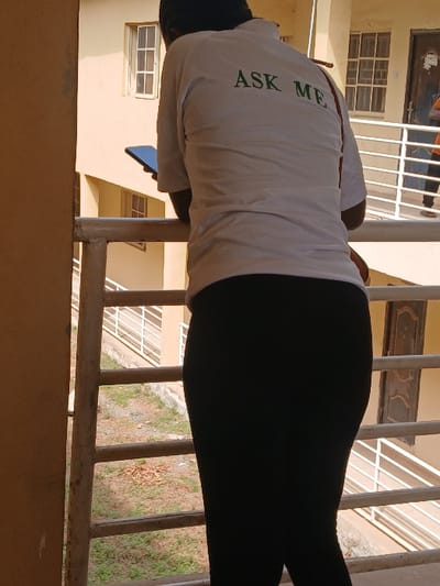 An outdoor area within a building complex in Ugi Majowel, Nigeria, is depicted under natural daylight. The foreground is dominated by a person viewed from the back, leaning forward over a white painted metal railing. This individual wears a white short-sleeved collared shirt with "ASK ME" printed in green capital letters across the upper back, and black leggings or tight-fitting trousers. They hold a dark-colored smartphone in their raised right hand. Below the railing, a patch of ground with sparse grass and scattered dry leaves is visible. Across this ground area, a light-colored multi-story building stands with visible multiple-pane windows and a dark brown door on its lower level. A second white metal railing defines a walkway on the lower level of the background building. A second individual is partially visible on this lower-level walkway near the dark brown door, wearing light-colored clothing.