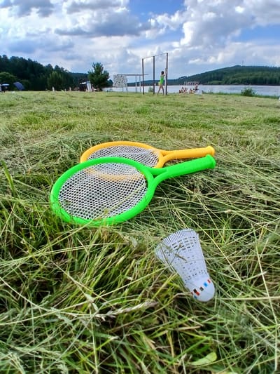 In the foreground of a grassy outdoor scene, two plastic badminton rackets and a shuttlecock lie on uncut grass. One racket is green and white, the other is yellow and white. The shuttlecock is white with colored accents at its base. In the mid-ground, a person in a green outfit stands near a rectangular structure. Several other people are seated on the grass further back. The background features a body of water, a tree-lined hill, and a cloudy sky. The scene appears to be a recreational area, possibly a park or waterfront in Revda, Russia.