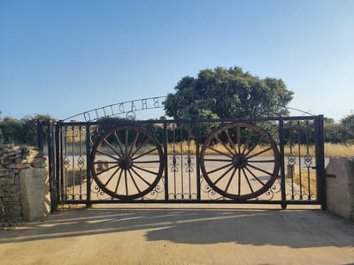 A wrought iron gate, decorated with two wagon wheels and the town name "SOBRADILLO" in ornate lettering, stands open under a clear blue sky. The gate is flanked by two rough-hewn stone pillars, one to the left with a small stacked stone wall adjacent. Behind the gate, a paved path leads into a landscape with dry grass, scrub brush, and a large, leafy green tree. Sunlight casts long shadows across the path in front of the gate. The scene is situated in the rural landscape of Sobradillo, Spain.