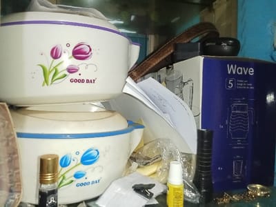This scene is situated in the Inxuba Yethemba Local Municipality, South Africa. The image displays a cluttered indoor surface, likely a shelf. Centrally, two white, octagonal, insulated food containers are stacked. The upper container features a purple rim and a graphic of three purple tulips with green stems, accompanied by the text "GOOD DAY" in black. The lower container has a blue rim and a graphic of blue tulips with green stems, also bearing the text "GOOD DAY." To the right, a vertical blue cardboard box labeled "Wave" displays an illustration of a jug and four glasses, with accompanying text indicating a "Juego de jarra." A brown, leather-textured strap is draped over the top food container and the blue box. Miscellaneous objects are scattered across the foreground and around the primary items: a small, golden-capped roll-on bottle stands to the left of the lower food container; loose white papers are present; a black cylindrical object, resembling a flashlight, stands upright near the blue box; a yellow plastic squeeze bottle with a white cap is on the surface; a transparent plastic bag contains indistinct items; a small black clip rests on a piece of paper; and a gold-colored, ornate metallic object lies to the far right. A small black, oval-shaped case or container is partially visible above the blue box, obscured by the brown strap. The background is an indistinct, uniform surface.
