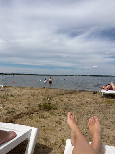 The image depicts a section of a sandy beach on a cloudy day, likely at the New Kremenkul' reservoir in the city of New Kremenkul', Russia. In the foreground, the legs and feet of a person are visible, resting on a white lounge chair. The sand is sparsely covered with dry grass and small plants. In the middle ground, a body of water, the reservoir, stretches across the frame. Several people are present in the water. Two individuals, a man and a woman, are standing partially submerged, with the woman wearing a headscarf. Further in the water, additional figures can be seen. Orange buoys are scattered across the water's surface, marking a swimming area. On the right side of the frame, another person is lying on a pink-cushioned lounge chair, also facing the water. The background shows a distant shoreline with buildings and trees under a sky filled with scattered clouds.