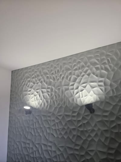 The image shows a wall with a textured, geometric pattern. Two spotlights are mounted on the wall, illuminating the texture. One spotlight is on the left, and the other is on the right. The wall is painted a neutral gray color, and the ceiling is white. The scene appears to be indoors, in a room. The image is a photograph taken from a slightly upward angle.