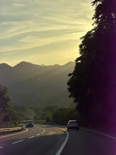 The image shows a winding road in Gospoština, Montenegro, bordered by trees and a mountain range in the background. Two cars are visible on the road. The sky is light with scattered clouds.