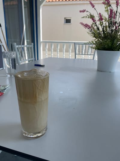 A tall glass of iced coffee sits on a white table outside a building in Skala Avdiron, Greece. The coffee is light brown and topped with a frothy layer. To the left of the coffee is a glass of water and a packet of something. Behind the table, there is a white railing and a building with a red-tiled roof. A potted plant with purple flowers sits to the right of the coffee.
