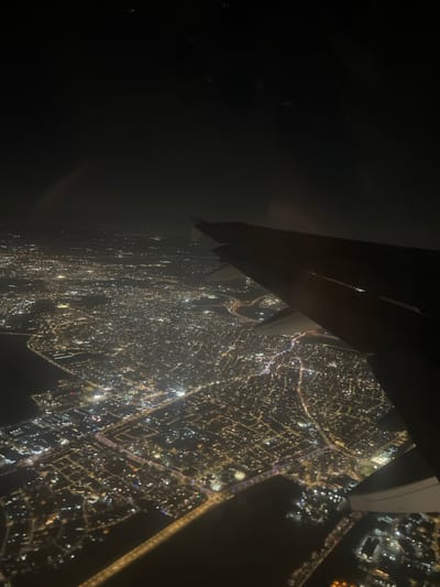An aerial photograph, captured at night from an aircraft window, depicts the illuminated urban expanse of Yehud Monosson, Israel. The dark wing of the aircraft occupies the right foreground and mid-ground, showing its structure against the brightly lit cityscape below. The city is characterized by a dense, intricate network of golden and white artificial lights delineating streets, buildings, and urban infrastructure. Dark, unlit areas, consistent with water bodies, are visible bordering the city lights on the left and interspersed throughout the landscape. The sky above is dark and featureless. No individuals are visible. The scene captures the city's nocturnal illumination from an elevated perspective during flight.