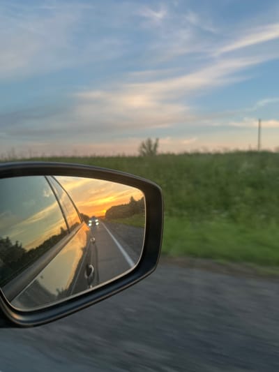 The image is a photograph taken from the perspective of a car driving on a road in Verkhnechusovskie Gorodki, Russia. The photo is framed by the driver's side mirror, which reflects the road behind the car. In the mirror, a sunset is visible, casting warm orange and yellow hues across the sky and reflecting on the car's surface. Two headlights from another vehicle are visible in the distance on the road. The surrounding environment appears to be a rural landscape with green fields and a cloudy sky with streaks of pink and blue. The road surface is dark asphalt.