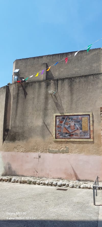 The image shows a building facade in Canals, Spain under a bright blue sky. A string of colorful triangular pennants hangs diagonally across the left side of the frame. The building's wall is predominantly gray, with a section of pink on the right. Affixed to the gray wall is a rectangular tiled artwork depicting an aerial view of a cityscape. A small light fixture is visible near the bottom left of the gray wall. A narrow sidewalk runs along the right edge, its surface a mottled gray concrete.