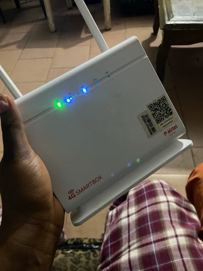 An eye-level, medium shot captures a person's dark-skinned hand holding a white Airtel 4G Smartbox router in a dimly lit room. The router is positioned at an upward angle, with its indicator lights illuminated. From left to right, the lights are green (SYS), blue (RF), blue (WIFI), blue (ETH), and blue (NET). A QR code sticker and a barcode sticker are affixed to the top right of the router. The text "4G SMARTBOX" is visible in red on the front of the router. The background is out of focus, showing tiled flooring and furniture. The scene is located in Orodo, Nigeria.