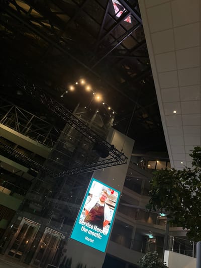 View looking upwards inside a multi-story building. A large screen displays "Service Hero of the month: Martin!" with a picture of a man. Steel support structures are visible, along with rows of lights. The architecture has a modern, industrial feel with many windows. Tree branches are seen near the top of the frame. Part of a white ceiling with recessed lights is also visible.