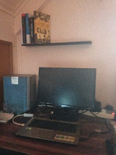 The image depicts an indoor workstation setup within a room in the city of Dogsenang Gorphoog, Bhutan. A dark wooden desk occupies the foreground, supporting a dark flat-screen monitor positioned centrally. In front of the monitor, a silver-grey laptop with an open lid rests on the desk, its keyboard and touchpad visible, along with multiple circular decals on the palm rest. To the left of the monitor, a dark blue-grey rectangular unit, possibly a speaker or computer tower, stands upright, displaying a light-colored label on its upper-front surface. A white cable with a connector extends from the left side of the laptop. A white keyboard is partially visible on the far left edge of the desk. To the right of the monitor, a white power strip with an illuminated red indicator light is visible, connected by dark cables.
Above the desk, a narrow dark shelf is mounted on a light-colored, textured wall. Three books are arranged on the shelf: two with dark spines on the left, and one with a yellowish-tan cover featuring text and photographic elements on the right. A reddish-brown wooden door frame or door is partially visible on the far left of the background. No individuals are present in the scene.