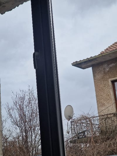 The image depicts an exterior view through a window, captured in the city of Yulievo, Bulgaria. In the immediate foreground on the left, a dark-colored window frame with a black handle is partially visible. A rough, grey concrete edge is present in the top-left corner. The sky is overcast and light grey. In the midground, several bare deciduous trees or bushes with exposed branches are visible. To the right, a section of a building is present. This building features a textured, light brown or beige facade, likely stucco, and a terracotta-tiled roof with a concrete eave and a rain gutter. A balcony with a black ornate metal railing is situated on this building, heavily covered by dried, tangled vines or leafless creepers. A white parabolic satellite dish is mounted on a pole near the balcony. A dark-framed window or door is partially visible on the building's facade above the balcony. No individuals are present in the scene.