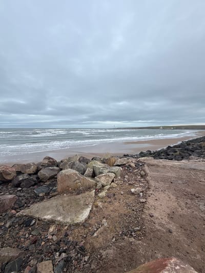 The image displays a coastal scene in Montrose, United Kingdom, under a uniform overcast, light grey sky. In the foreground, a rough, dark brown and reddish-hued dirt pathway occupies the right, transitioning into an area of scattered dark and multi-colored small rocks and debris. To the left, a substantial cluster of large, irregular boulders, primarily grey and brown, forms a rock barrier adjacent to the ocean. A broken, irregularly shaped light grey concrete slab with visible white edges is present on the ground amidst the small rocks in the immediate foreground. Beyond these elements, a wide sandy beach is visible, with numerous waves breaking and receding, displaying white foam. The ocean water appears dark grey-green. In the distant background, a white lighthouse or beacon stands at the end of a dark landmass or breakwater extending into the sea from the right. Further along this distant coastline to the far right, a low, green headland is observable, featuring several small, white structures at its base. No individuals or specific human actions are present within the frame.