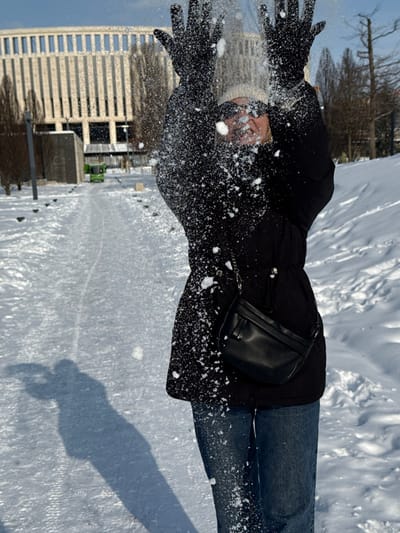 The scene depicts a sunny winter day in an urban park or public pathway within Plodorodny, Russia. A single individual, dressed in a black winter coat, blue jeans, black gloves, and a light-colored beanie, is captured in motion. The individual's arms are raised overhead, hands open, actively releasing a quantity of snow upwards. Numerous snowflakes and small snow fragments are suspended in the air, primarily obscuring the person's upper face and torso. A black cross-body bag with a chain strap is worn across their chest. The ground is covered in uniform white snow, traversed by a visible, compacted path exhibiting areas of ice and snow. Long shadows, including that of the individual, are cast across the snow, indicating clear weather and direct sunlight. In the background, a prominent multi-story building with a light-colored, vertically-oriented facade and multiple windows is visible. Bare deciduous trees line the path on both sides. Further along the path, a small, bright green utility vehicle is discernible. The environment is consistent with a maintained public space during winter conditions.