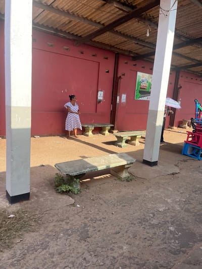 The image depicts an outdoor scene at a bus stand in Moratuwa, Sri Lanka. A woman in a patterned dress stands by a red wall, facing left and looking away from the camera. Several concrete benches are present in the foreground and midground, some with visible weathering and small plants growing around them. In the background, a sign with the text "BIO Clean Railway" is visible on the red wall. To the right, a person holds a colorful umbrella, and some exercise equipment is stacked. The ground is a mixture of dirt and concrete. The architecture suggests an open-air structure with a corrugated roof supported by wooden beams and prominent white pillars. The overall impression is a functional, somewhat worn, public space.