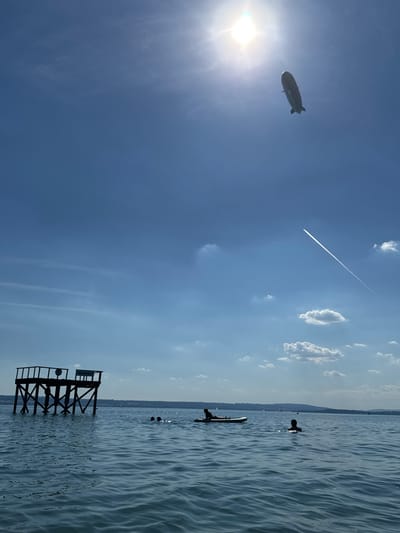 This is a vertical, daytime, outdoor, wide shot of a lake. The sun is shining brightly in the upper left corner, with a zeppelin flying in the sky. A contrail from a jet can be seen diagonally across the sky. In the water, two people are on stand-up paddleboards, one of whom is partially submerged. Further back, a few other figures are in the water. In the foreground, a wooden diving platform stands in the water. The overall scene suggests a warm, sunny day at a lake, possibly a recreational area. The water is calm with gentle ripples. The horizon shows a distant shoreline with hills. There is no visible text.
