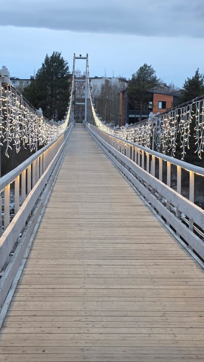 The image shows a long, wooden pedestrian suspension bridge in Kandalaksha, Russia. The bridge deck is made of vertical wooden planks. White, ladder-style railings flank both sides of the bridge. Decorations of small, white lights are strung along the railings. In the background, buildings and trees are visible. The sky is light blue and overcast. The perspective of the image suggests it was taken from the bridge, looking straight down its length.