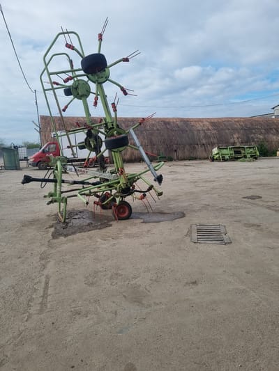 The image shows a green agricultural hay rake implement parked on a concrete surface. In the background is a large, curved, corrugated metal building and a green agricultural machine. Further back, parts of a red truck and white containers are visible. The sky with scattered clouds is in the top left corner. A drainage grate is on the concrete surface. The location is Ovcharovo, Bulgaria.
