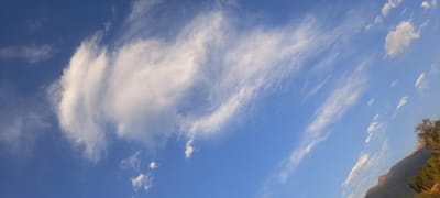 The image displays a vast, clear blue sky with scattered white clouds. A large, swirling cloud formation is visible on the left side of the frame, with wispy cirrus clouds stretching across the sky towards the right. In the far right of the image, the upper slopes of a mountainous terrain covered in vegetation are visible, suggesting a natural, possibly rural or undeveloped, environment. The overall impression is of a bright, open sky with a backdrop of natural scenery. The location is identified as Bash-Kaiyndy, Kyrgyzstan. There are no people, animals, or man-made objects clearly discernible in the image besides the implied natural landscape. The lighting suggests it is daytime, likely with the sun in the sky, casting shadows or highlighting the clouds and mountain.