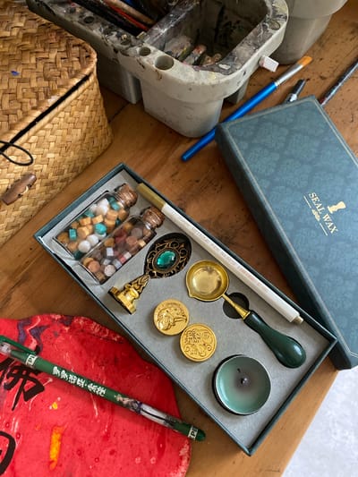 The image displays a collection of art and craft supplies arranged on a wooden surface. In the foreground, a red mat with black and purple abstract ink markings is visible, upon which a green pen with Chinese characters and an illustration is resting. To the right of the mat is a dark green box containing a seal wax set. The set includes two small glass bottles filled with colorful wax beads, a metallic spoon for melting wax, a wax stamp with a decorative emerald-green gem and golden handle, two round golden wax seals with intricate embossed designs, and a small green dish with a remnant of a burnt candle wick. A white metallic pen with text "GUANGDA METALLIC PEN No. 6510" is also placed within the box. Behind the seal wax box, a dark blue-green book or box with embossed patterns and the text "SEAL WAX" in gold lettering is positioned. Further in the background, a woven straw basket with a wooden toggle closure is on the left. To the right of the basket is a white plastic container holding various paintbrushes with colorful bristles, some of which are stained. A blue paintbrush and another brush are also visible near the container. The overall scene suggests a workspace dedicated to art or crafting, possibly calligraphy or wax sealing. The presence of Chinese characters on the pen and the specific style of the wax seals might indicate a connection to Chinese culture or artistic traditions. The scene does not provide any visual cues to determine its specific location in Dujiangyan City, China; it is a generalized depiction of an artist's or crafter's setup.
