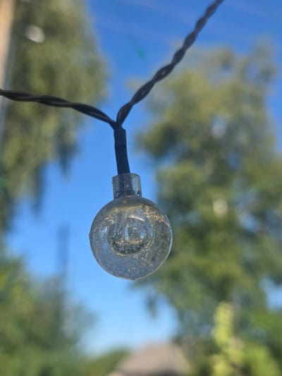 The image is a close-up, slightly low-angle shot of a single clear glass lightbulb from a string of lights. The lightbulb is suspended by a black twisted wire and is slightly out of focus. Inside the bulb, there are visible air bubbles and possibly filament remnants, suggesting it is either unlit or a decorative bulb. The background is blurred, showing a bright blue sky with soft green foliage from trees. The light is coming from above and slightly to the right, casting subtle highlights on the bulb. The image evokes a feeling of an outdoor summer day, possibly in a backyard or park setting. While the image itself does not contain specific geographical markers, the provided location of Ходасаўскі сельскі Савет, Belarus, suggests this scene could be in a rural or suburban area within that region. There are no people or animals present in the image.