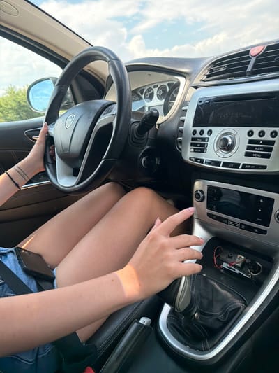 The image shows the interior of a car, taken from the perspective of a passenger. A person's legs, wearing denim shorts, are visible in the foreground, with their right hand on the gear shift. The car's dashboard, steering wheel, and center console are also visible. The overall impression is that of a person driving or being driven on a sunny day, with the sky and trees visible through the windshield and side window. The location is identified as Aleksandrovo, Bulgaria.