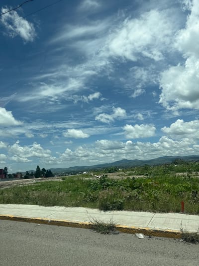 This is a landscape photograph taken in the city of San Francisco Totimehuacán, Mexico. The image captures a view of a semi-urban area with undeveloped land in the foreground and middle ground. The foreground consists of an asphalt road with a yellow-lined concrete curb and sidewalk. Beyond the sidewalk, there is a grassy area with some weeds and a small red marker stake. In the middle ground, there are some partially constructed buildings, trees, and more overgrown vegetation, indicating a developing or unfinished construction zone. Further in the distance, a town or city stretches across a valley, with a range of green hills forming the backdrop. The sky is a prominent feature, filled with a mix of white, fluffy cumulus clouds and wispy cirrus clouds against a bright blue sky. There are no people or animals visible in the image. The overall impression is one of a sunny day in a transitional landscape on the outskirts of a populated area.