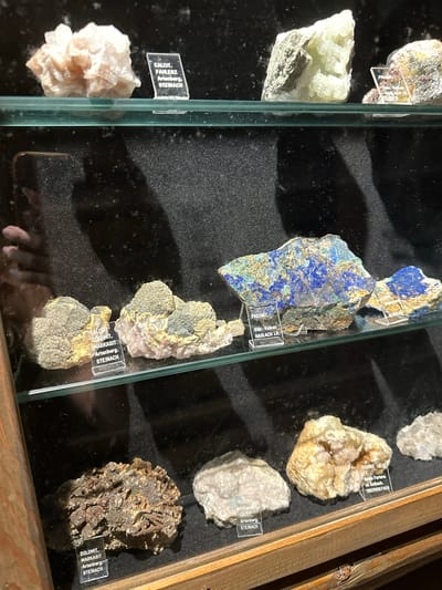 This media file captures a close-up view of a display case filled with various mineral specimens. The minerals are presented on a dark, textured surface, likely velvet, and illuminated from above. Each specimen is accompanied by a small, transparent label detailing its type and origin, such as "CALCIT, FAHLERZ Artenberg, STEINACH" and "FLUORIT Grube Fortuna Im Belbach, OBERWOLFACH."
The collection features a variety of rock types and colors, including pale green, blue, white, and earthy brown specimens. One prominent specimen in the center is a large piece of rock with striking blue and golden-brown veins, identified with labels like "PREHNIT Stbr. Vulkan HASLACH I.K." Other notable minerals include a cluster of pale pink, crystalline formations and a rough, dark brown mineral specimen. The display is housed within a wooden frame, suggesting it is part of a museum, collection, or shop. The lighting creates shadows and highlights, emphasizing the textures and facets of the minerals. The overall impression is one of a well-curated geological display. The provided location context of Schonach im Schwarzwald, Germany, suggests this is a local collection or exhibit of minerals from Germany.
