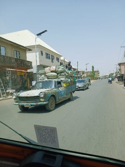 The image captures a daylight street scene in the city of Kungulu School, Zambia. An aged, heavily rusted light blue pickup truck, with significant paint deterioration, drives on a paved road. Its open bed is piled high with cargo, including various sacks and cardboard boxes. Two individuals are positioned on top of this cargo: one seated towards the front of the load, dressed in dark attire, and another standing or crouching further back, also in dark clothing. Behind the pickup, a silver sedan and other vehicles are visible in the distance, following the truck. On the left side of the road, two-story buildings are present; the nearest features a dark balcony railing, while an adjacent structure has a white metal roof and a prominent solar-powered street light. Buildings and utility poles line the right side of the street. The photograph is taken from inside a vehicle, evidenced by a visible dashboard section and a white rectangular card with a QR code affixed to the windshield in the foreground.