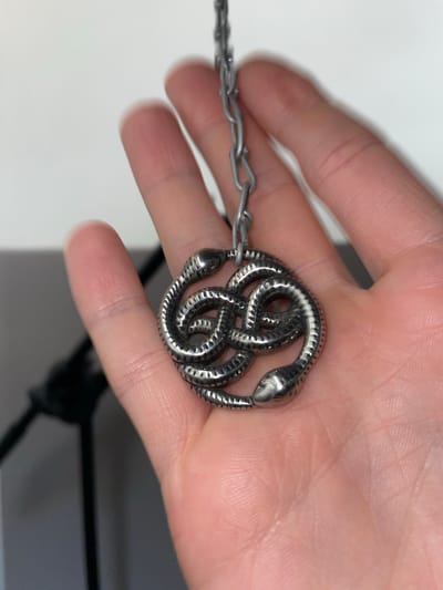 A human hand holds a silver-colored necklace featuring a pendant composed of intertwined snake figures. The necklace is suspended from the hand by a chain. The background is blurred, but hints of a white surface and a dark object are visible. There is no information given in the image about the location being La Lucila, Argentina.