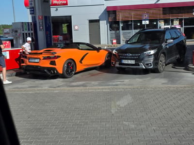 In Rotmanka, Poland, an orange Chevrolet Corvette convertible is parked at a gas station next to a black Subaru Outback SUV. The Corvette is positioned in front of the gas pumps, with its rear end facing the viewer. A person wearing a white cap and t-shirt stands near the rear of the Corvette, appearing to be interacting with it. The Subaru Outback is parked to the right of the Corvette, closer to the convenience store.
The gas station features prominent signage, including "ORLEN," "Myjnia" (car wash), and various fuel types like "verva 98," "verva 95," and "DIESEL." The convenience store section has advertisements displaying food items such as "bułkanapki" (sandwiches) and "śniadania" (breakfast). License plates are visible on both vehicles: "GDA 8811E" on the Corvette and "GD 5L 123" on the Subaru.
The scene appears to be taking place during the daytime, with clear sunlight casting shadows on the ground. The ground is paved with interlocking bricks. The overall atmosphere seems ordinary, with people conducting everyday activities at a gas station. No strong emotions are discernible from the subjects.